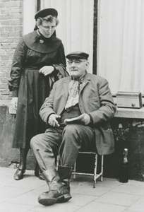 876923 Afbeelding van een Zuster Augustines die een praatje maakt met een oudere man die aan een onbekende straat zit, ...